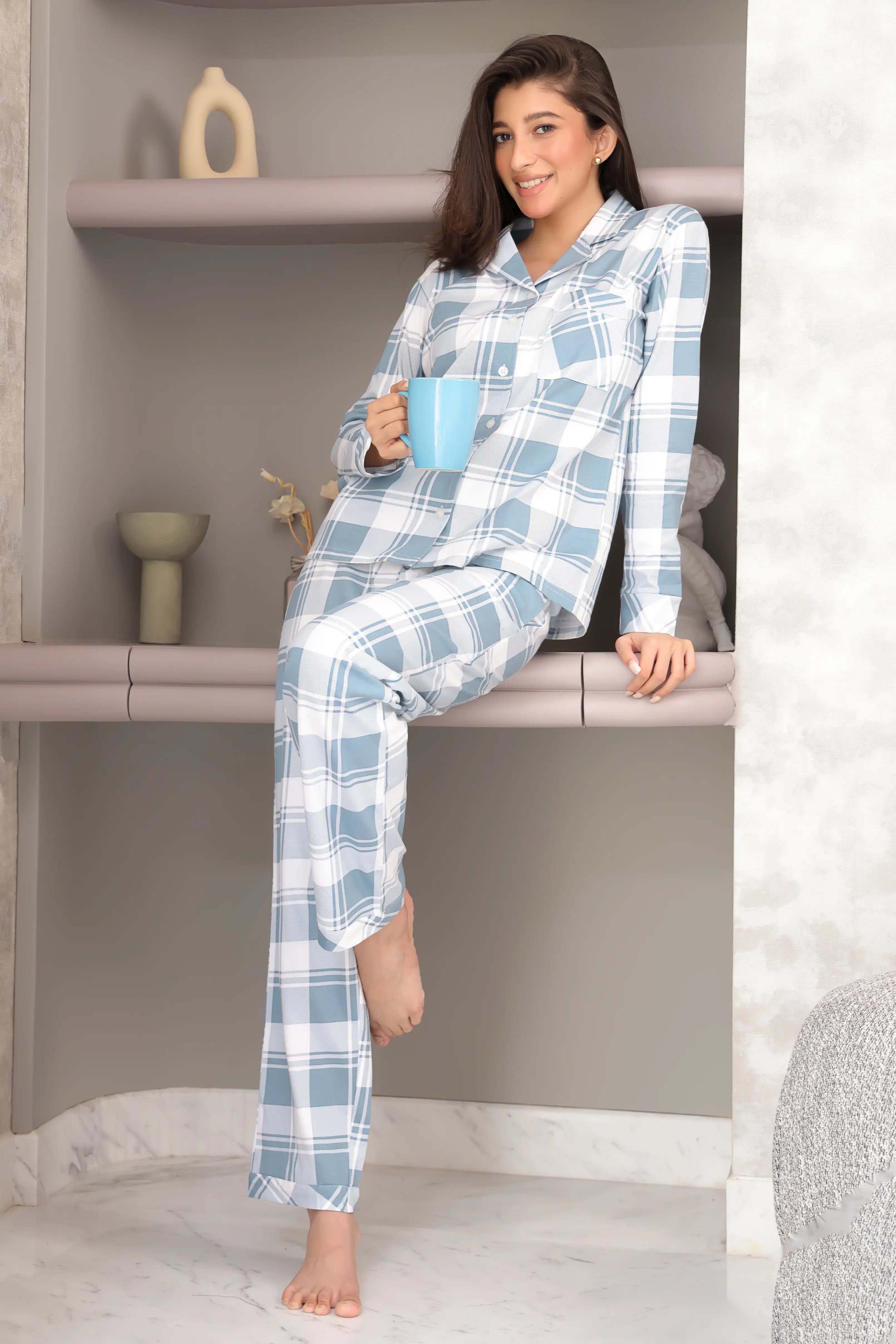 Woman in blue plaid pajamas holding a mug, sitting on a shelf.