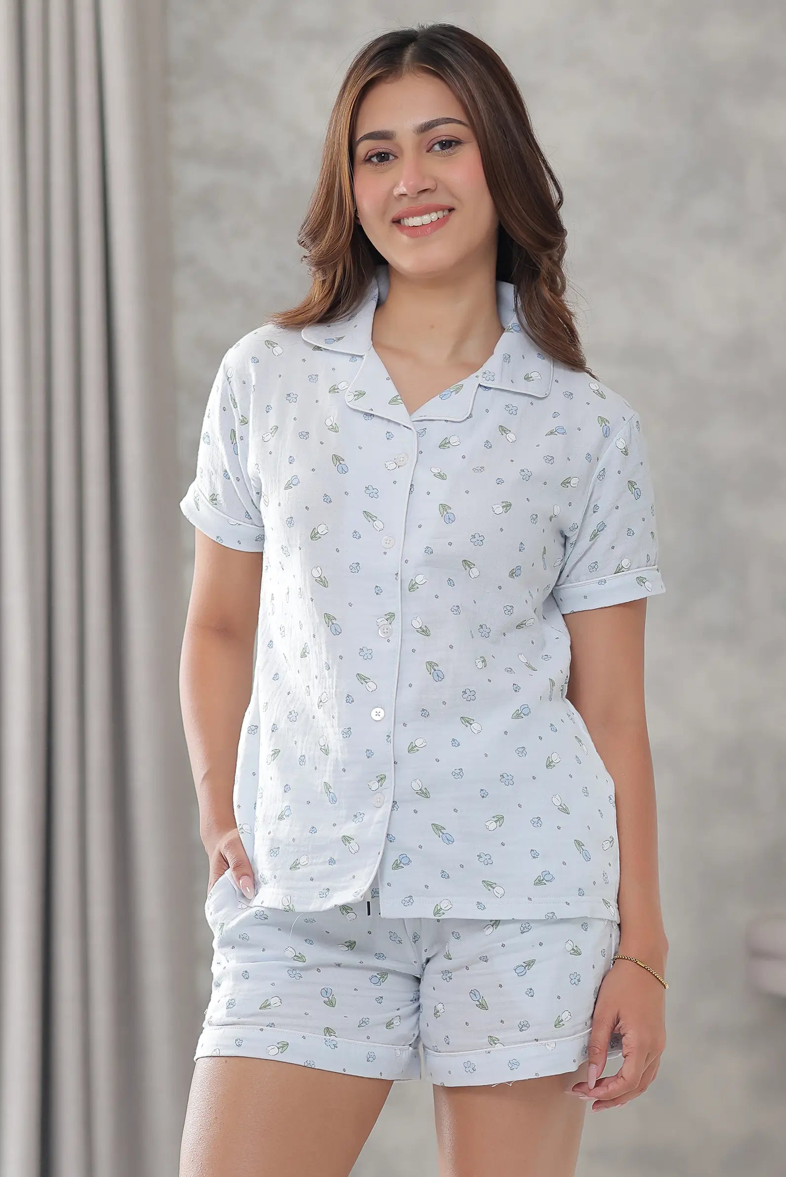 A woman modeling a two-piece shorts set consisting of a short-sleeve button-up shirt and matching shorts, both in a very light blue fabric with a small scattered floral print.