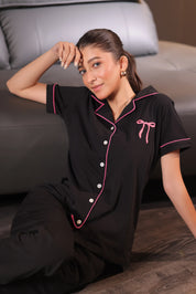 Woman wearing a black pajama set with pink accents sitting on a couch.