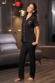 Woman wearing black embroidered pajamas with Pink piping in a living room setting.
