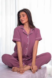 Woman wearing a purple pajama set with white piping sitting on a bed.