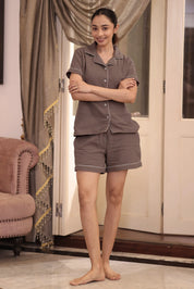 Woman wearing a brown shorts set standing in a room with curtains and a chair.