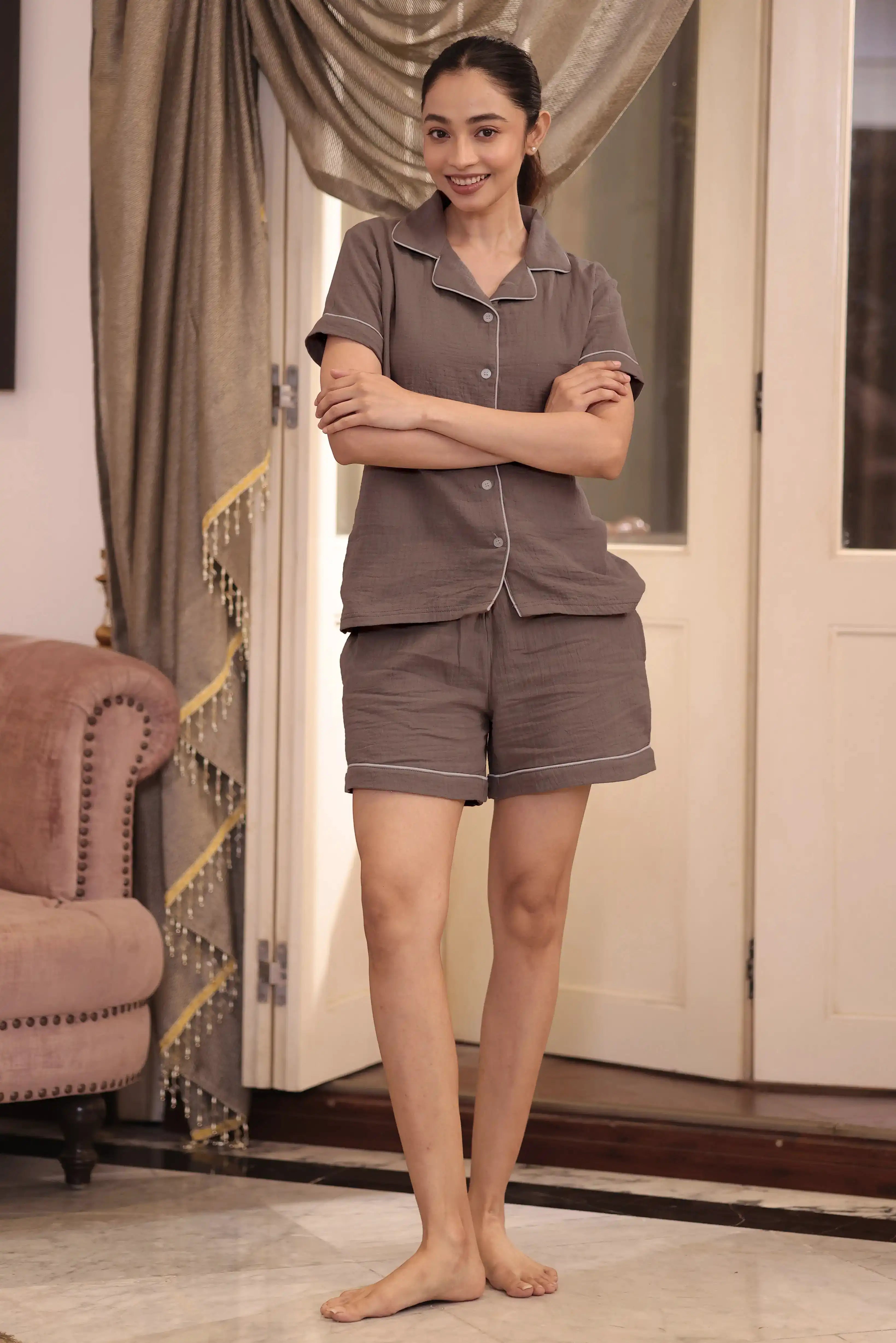 Woman wearing a brown shorts set standing in a room with curtains and a chair.