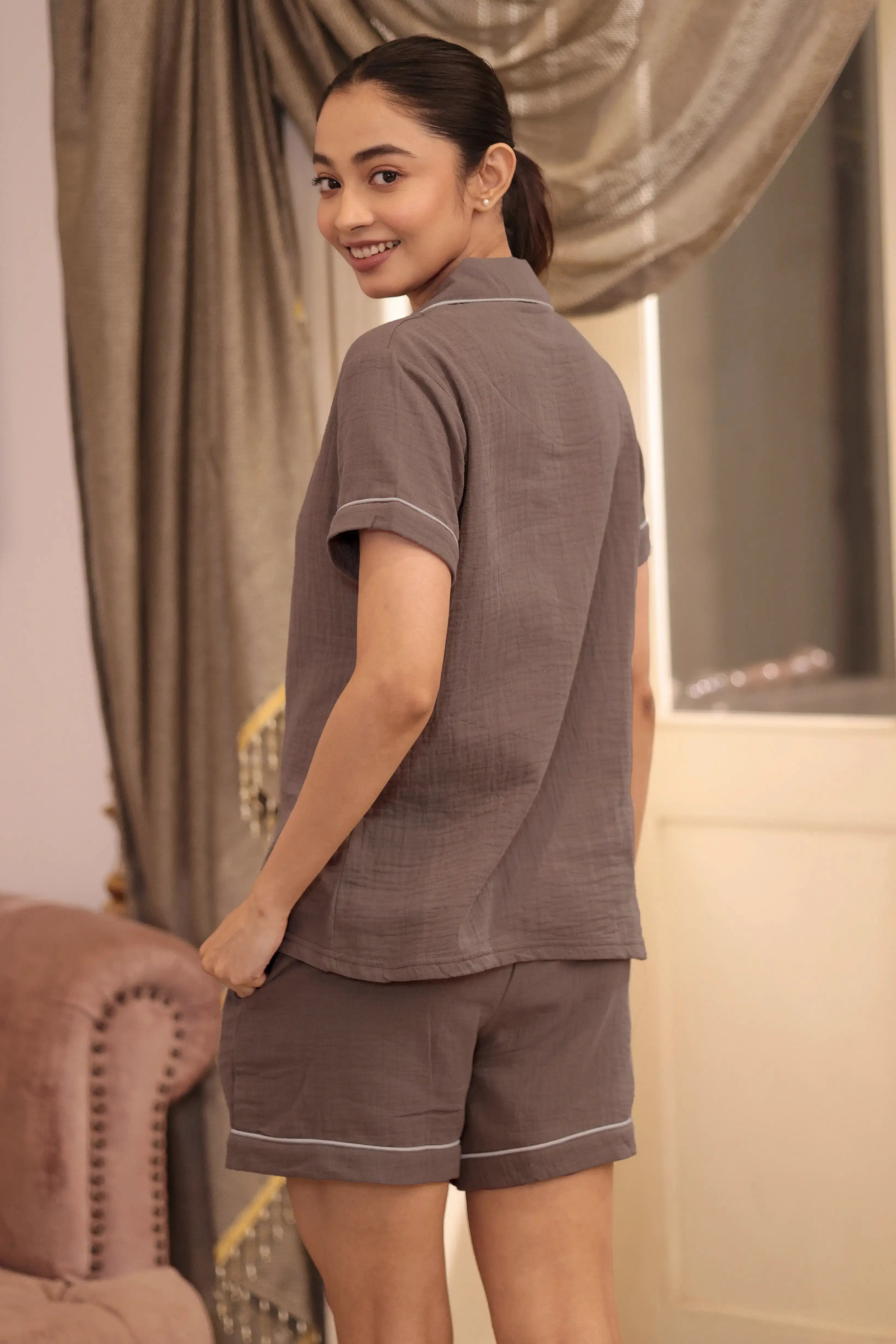 Woman wearing a brown shorts set in a room with curtains and a chair.