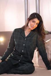 Woman wearing a green plaid pajama set sitting on a bed.