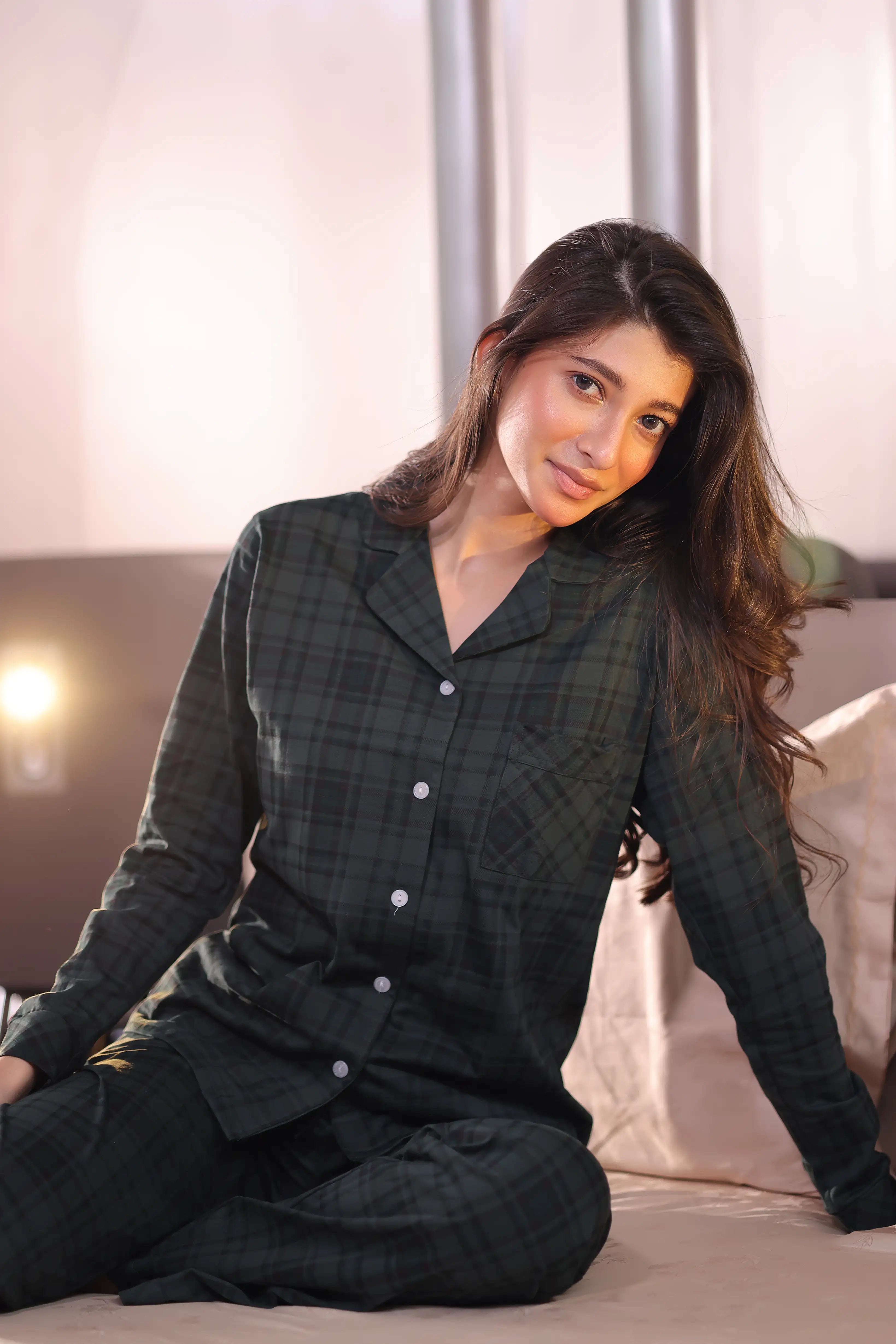 Woman wearing a green plaid pajama set sitting on a bed.