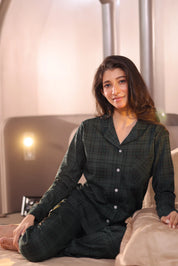 A woman wearing a dark green checkered outfit is sitting on a bed in a softly lit room.