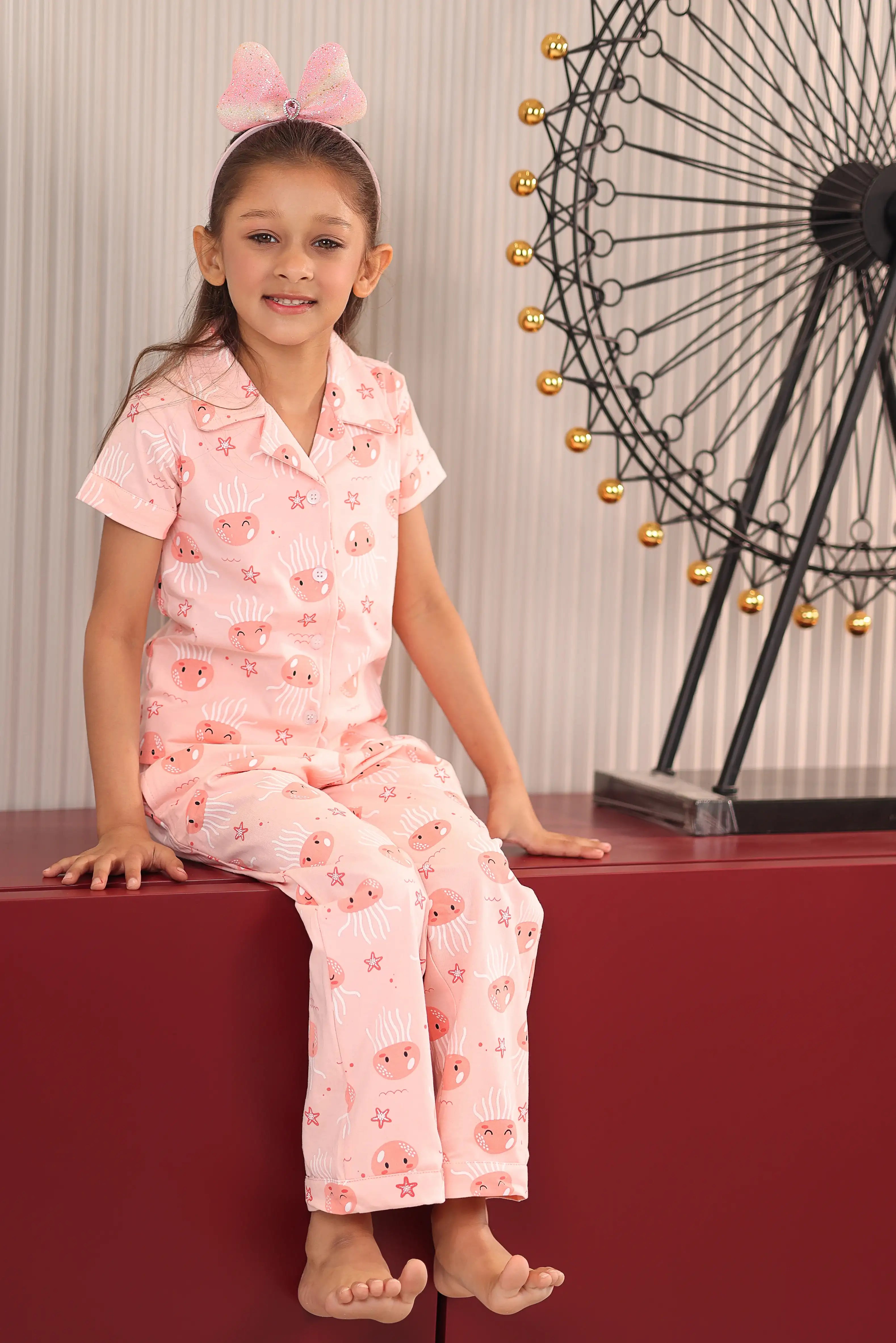 A girl in a pink pajama set with jellyfish print pattern, sitting on a red surface, ferris wheel in the background.