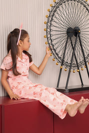 A girl in a pink print outfit is sitting on a red surface with a decorative wheel in the background.