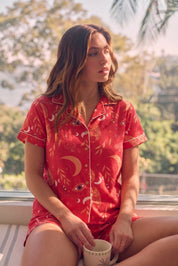 Woman wearing a red Shorts set with a pattern, sitting by a window.