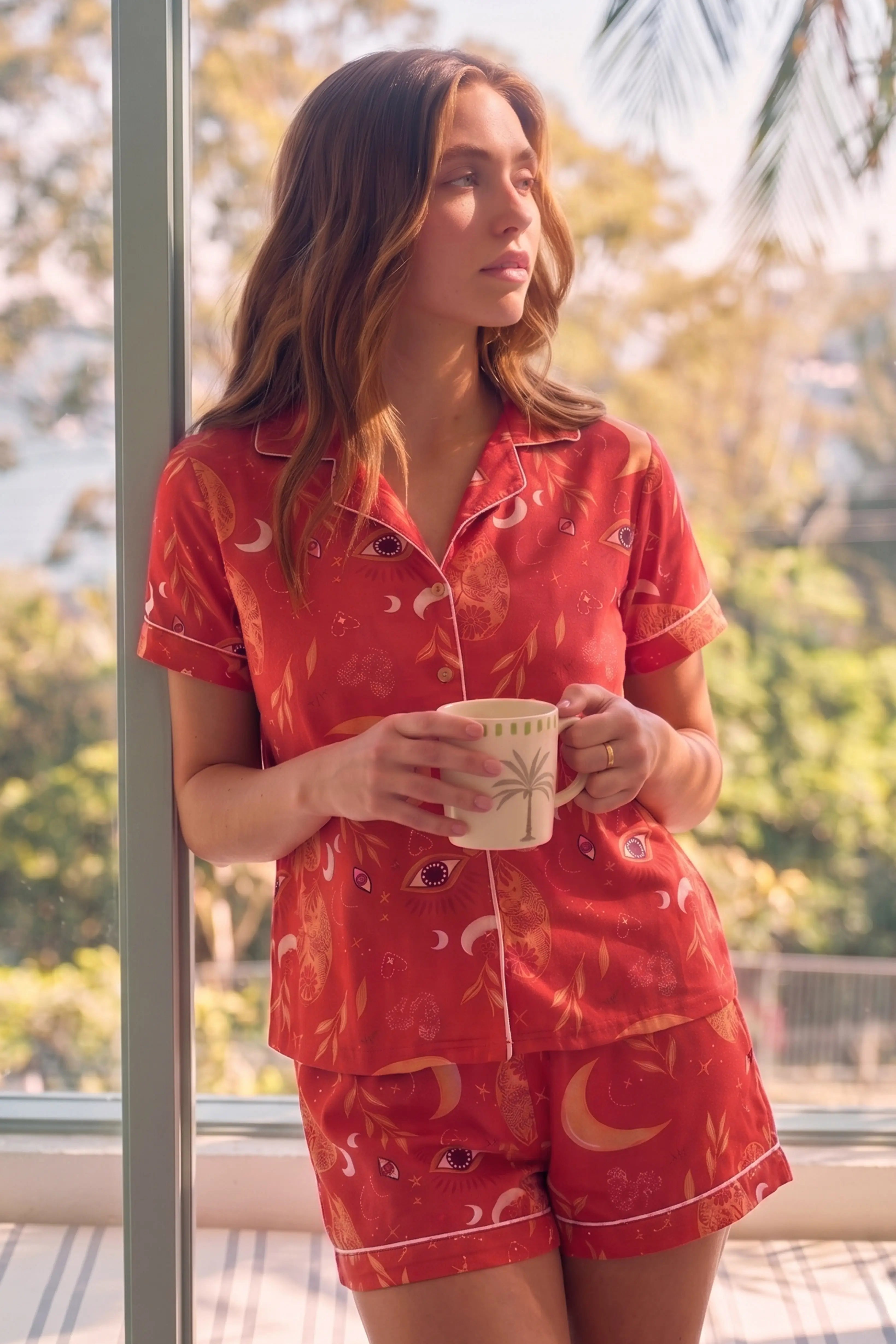 A woman in red shorts set holding a mug by a window with greenery outside