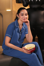 A woman in blue muslin pajamas holding a bowl of popcorn sitting on a couch.