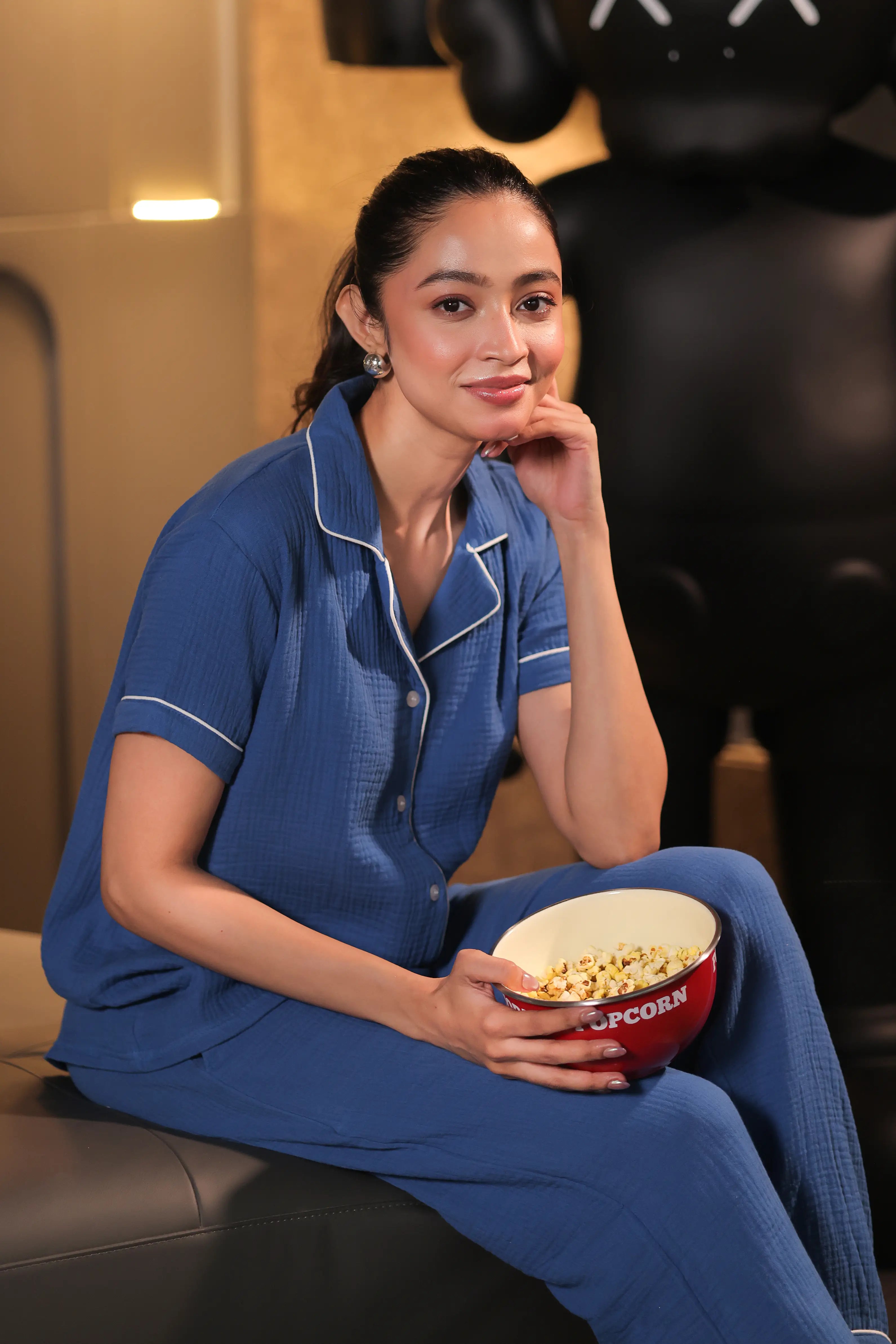 A woman in blue muslin pajamas holding a bowl of popcorn sitting on a couch.