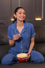 Woman in solid blue muslin pajamas holding a bowl of popcorn on a dark couch.