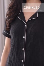 A close-up of a woman wearing a black crinkled short-sleeve Shorts top with white piping and white buttons. The text 'CRINKLED' is overlaid at the top.