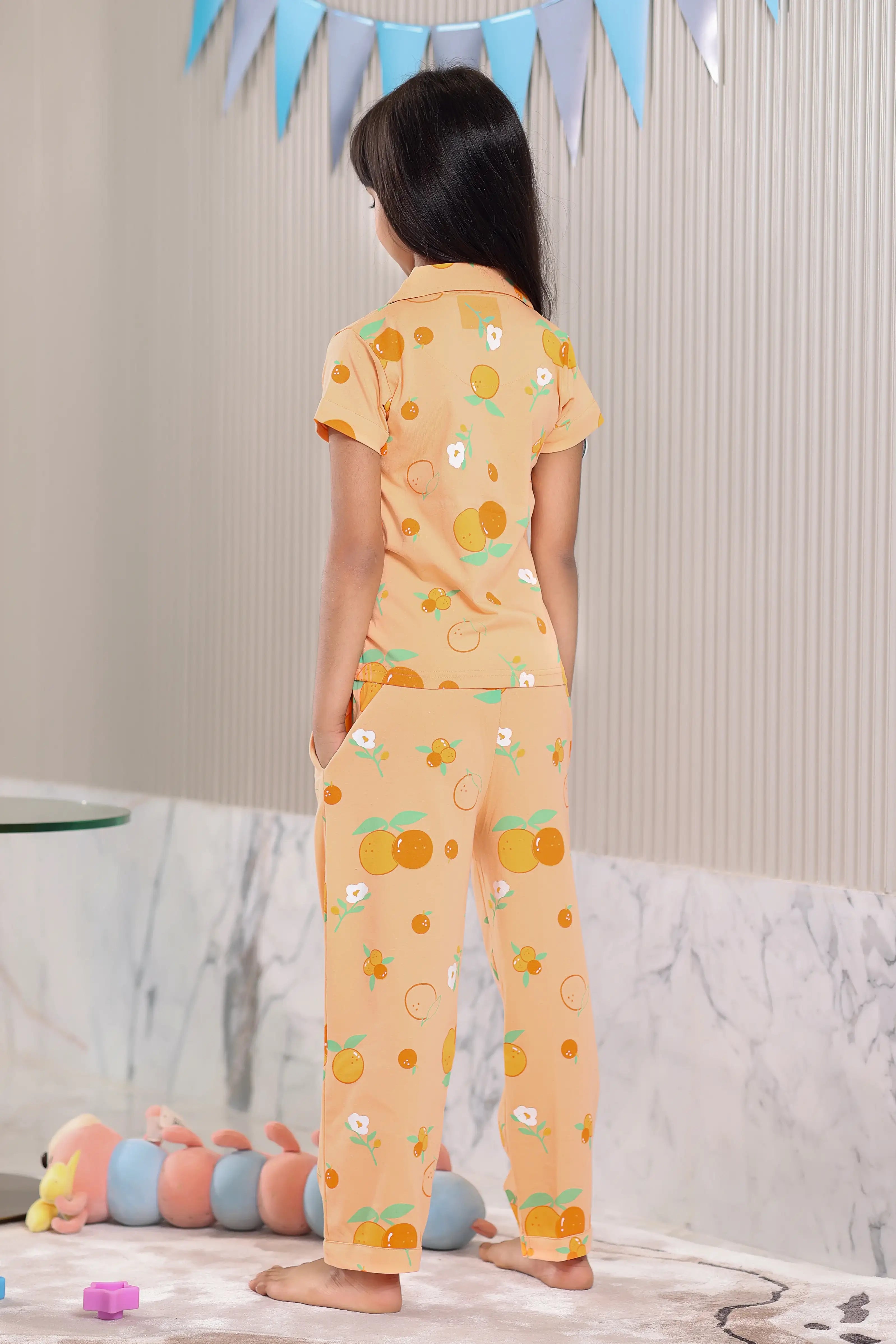 The girl wearing an orange-colored pajama set with an orange fruit pattern is in a room.