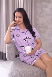 Woman wearing a purple shorts set with panda print, sitting on a gray chair holding a white mug.