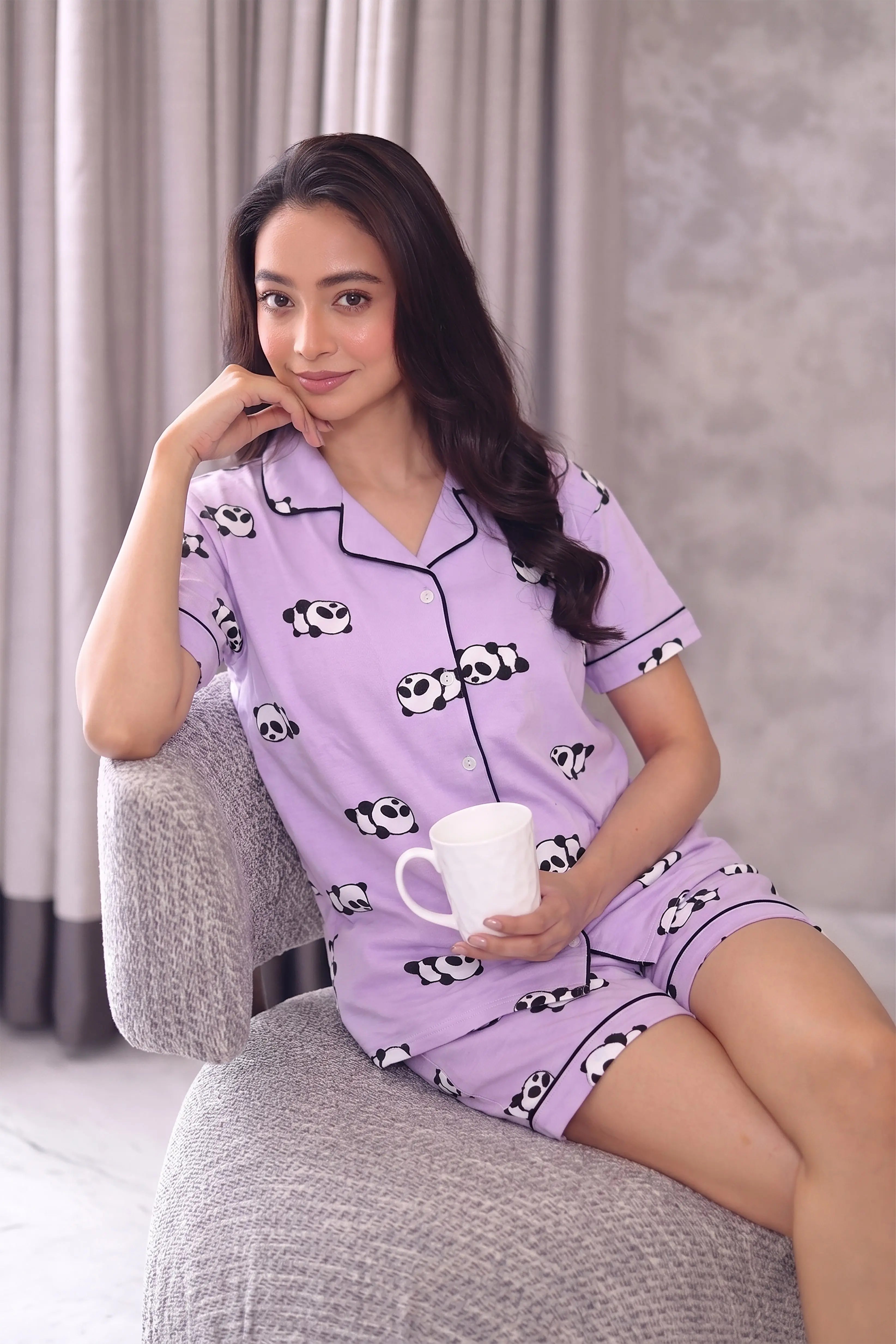 Woman wearing a purple shorts set with panda print, sitting on a gray chair holding a white mug.