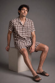 Man wearing a checkered shirt and shorts sitting on a block against a gray background