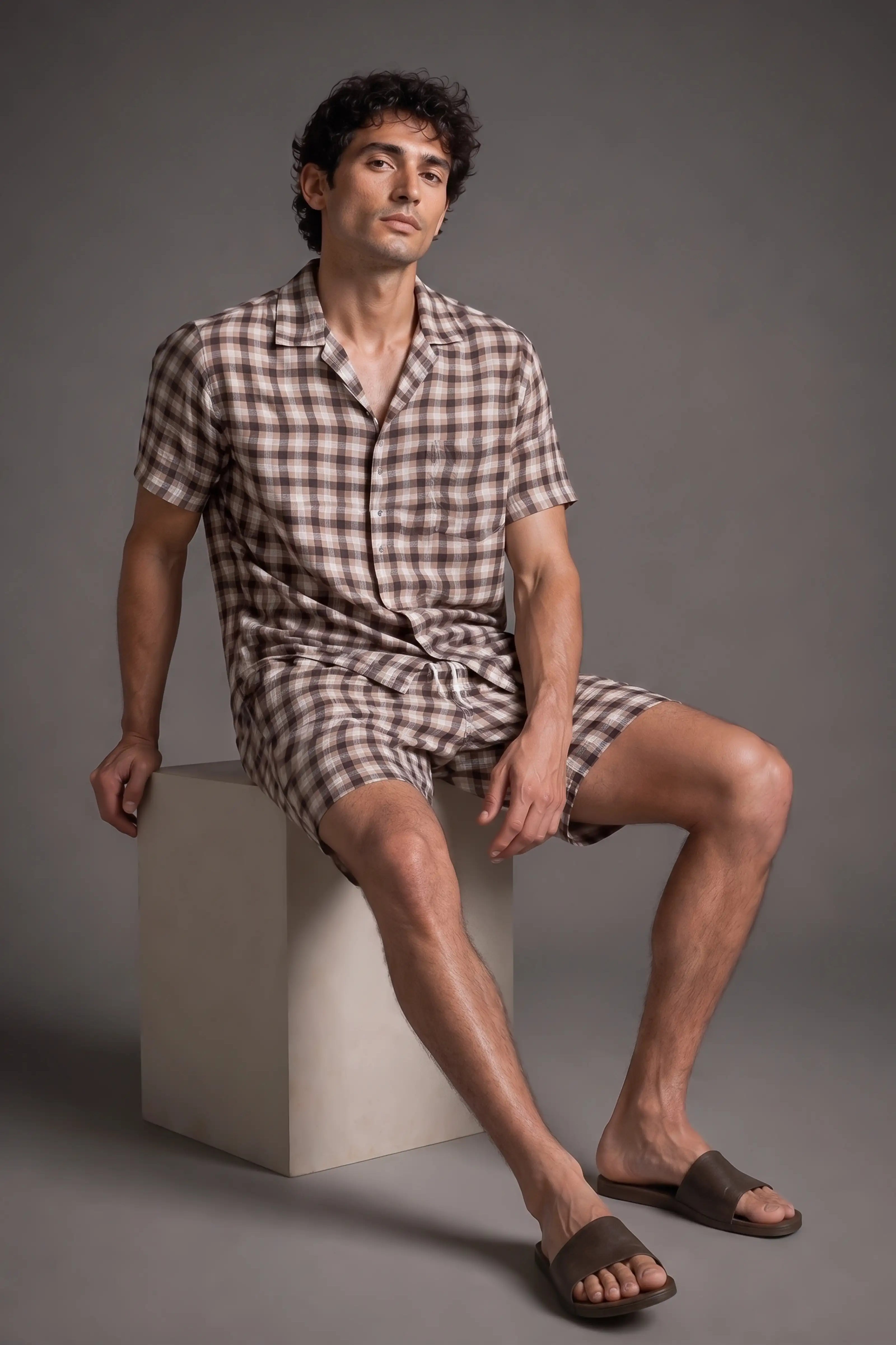 Man wearing a checkered shirt and shorts sitting on a block against a gray background
