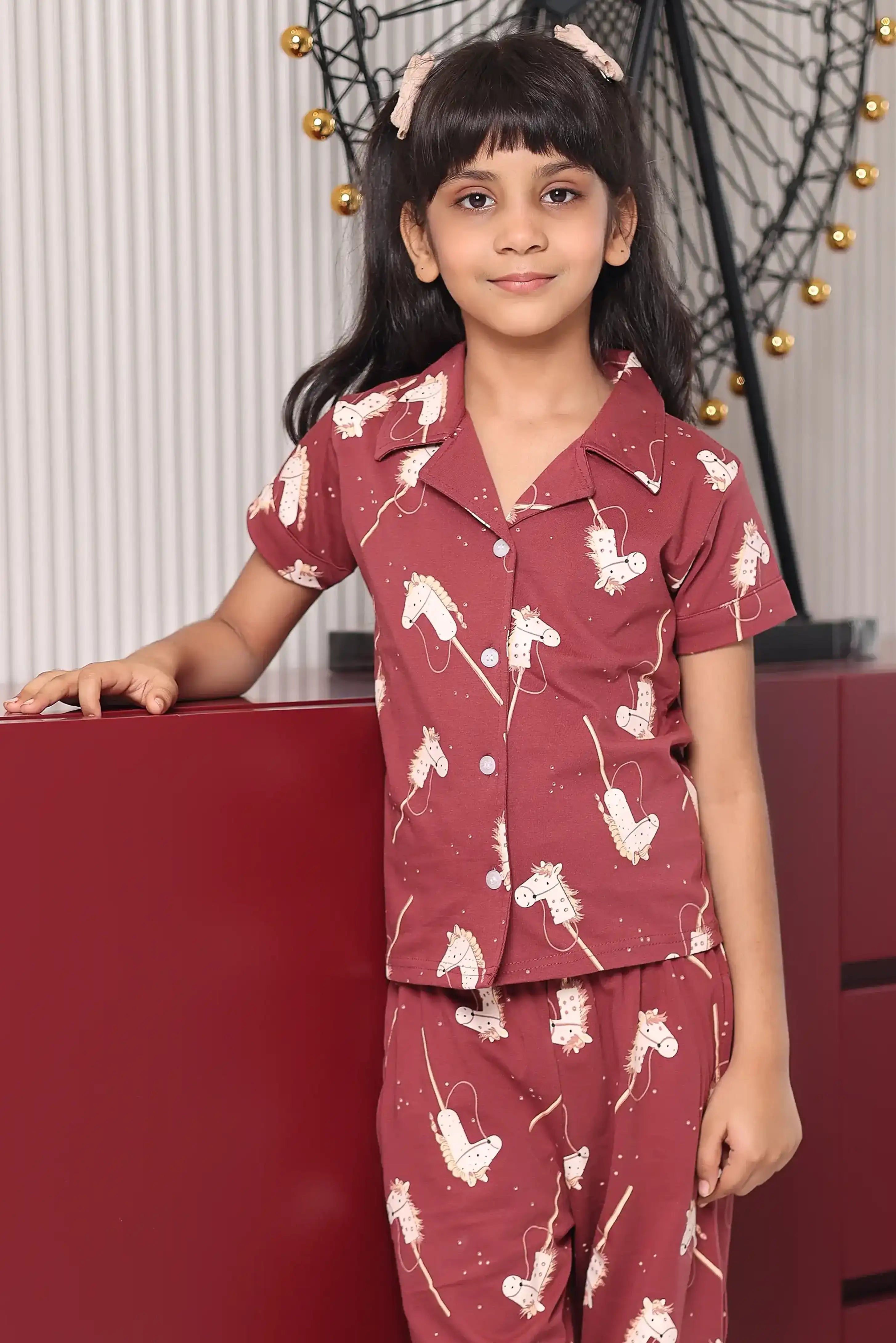 A girl wearing a maroon pony Print pajama set with white patterns, standing against a red background.