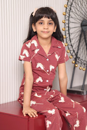A girl wearing a maroon pony print pajama set with a horse print. Girl sitting on a red surface.