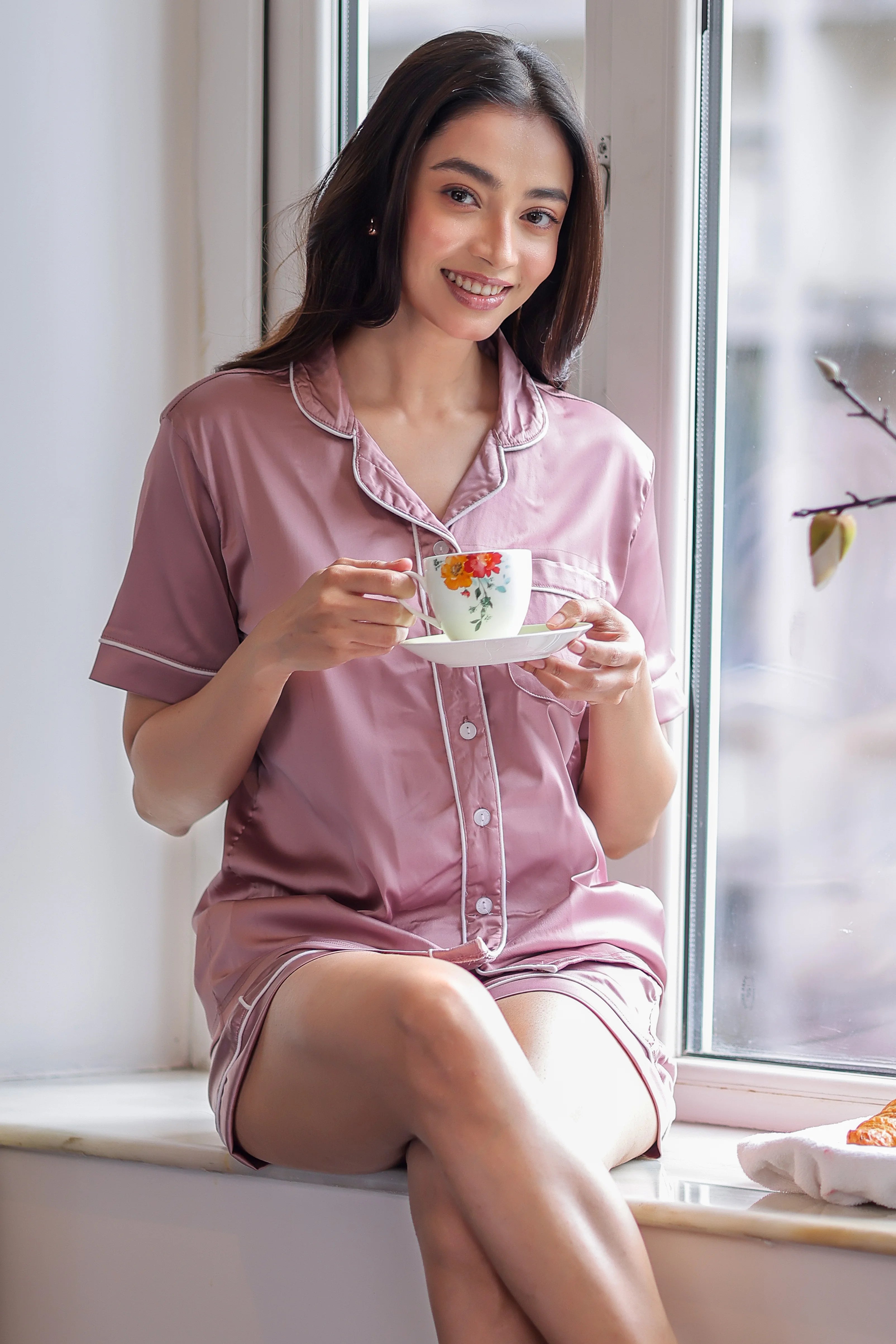 Woman in pink shorts set holding a floral teacup by a window