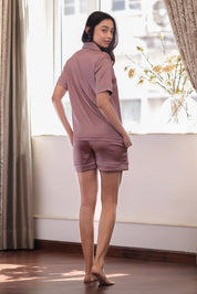Woman wearing a mauve shorts set standing in a room with curtains and flowers.