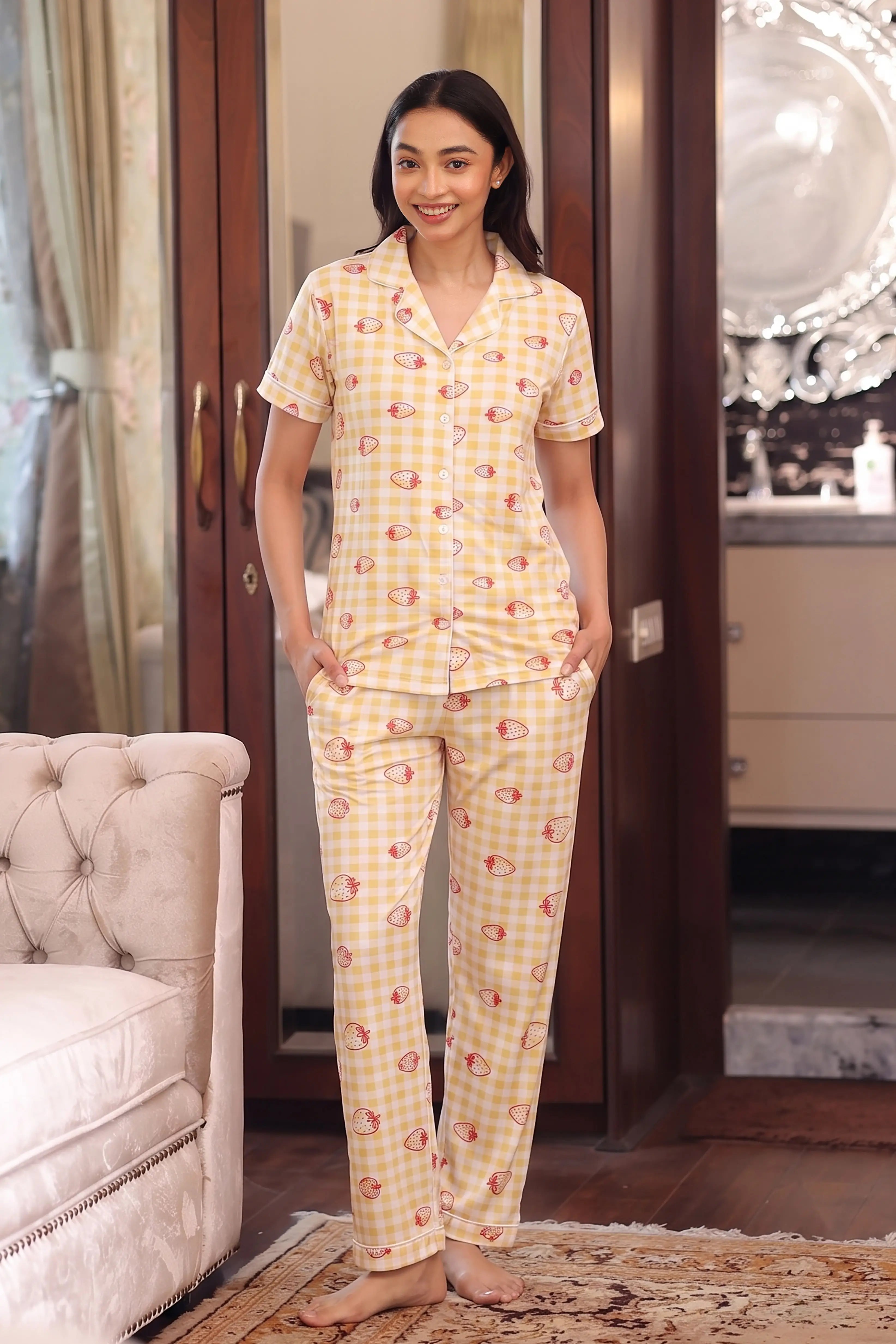 Woman wearing a yellow strawberry checkered pajama set in a room with a chair and decorative items.