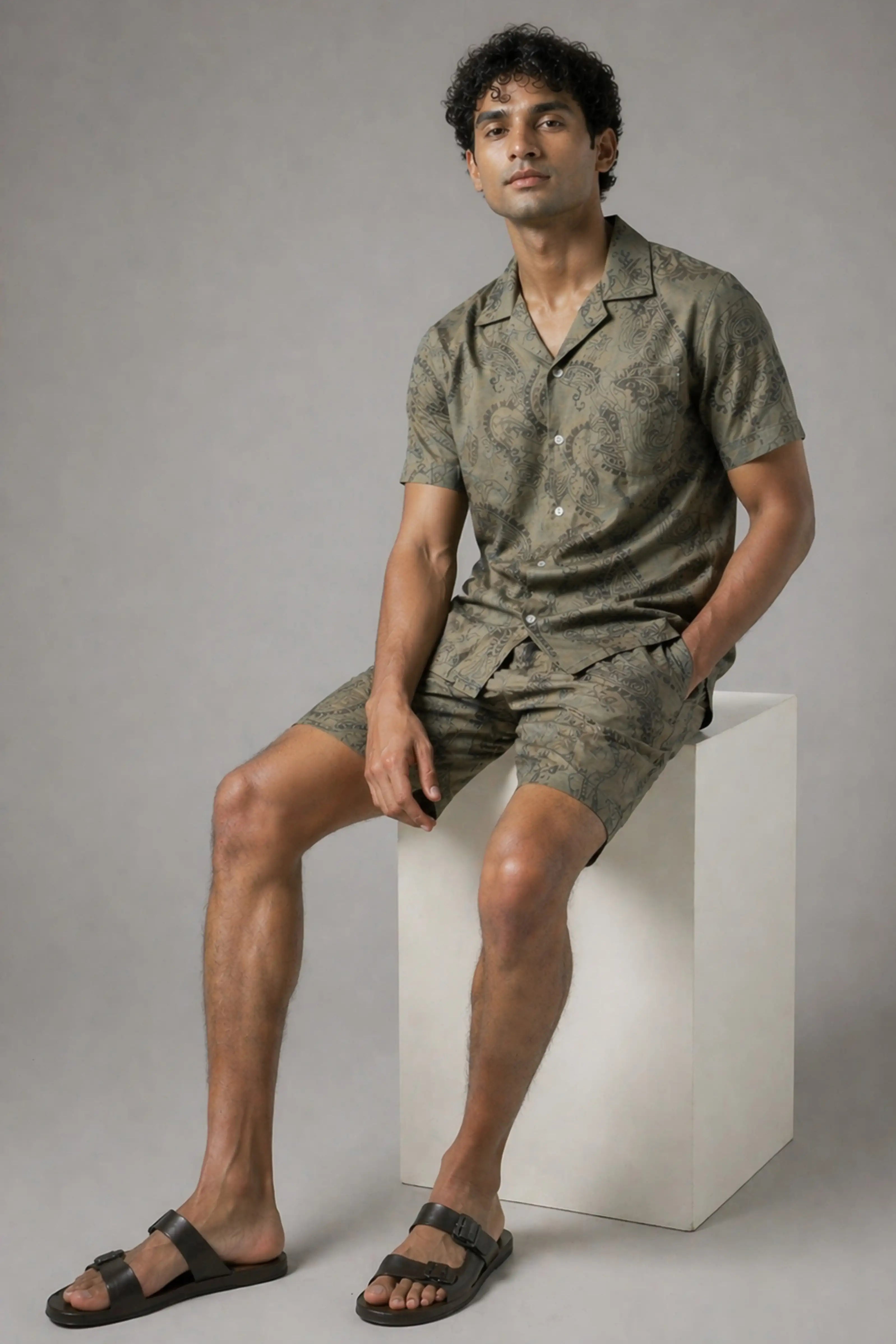 Man wearing a green patterned outfit sitting on a white block against a gray background