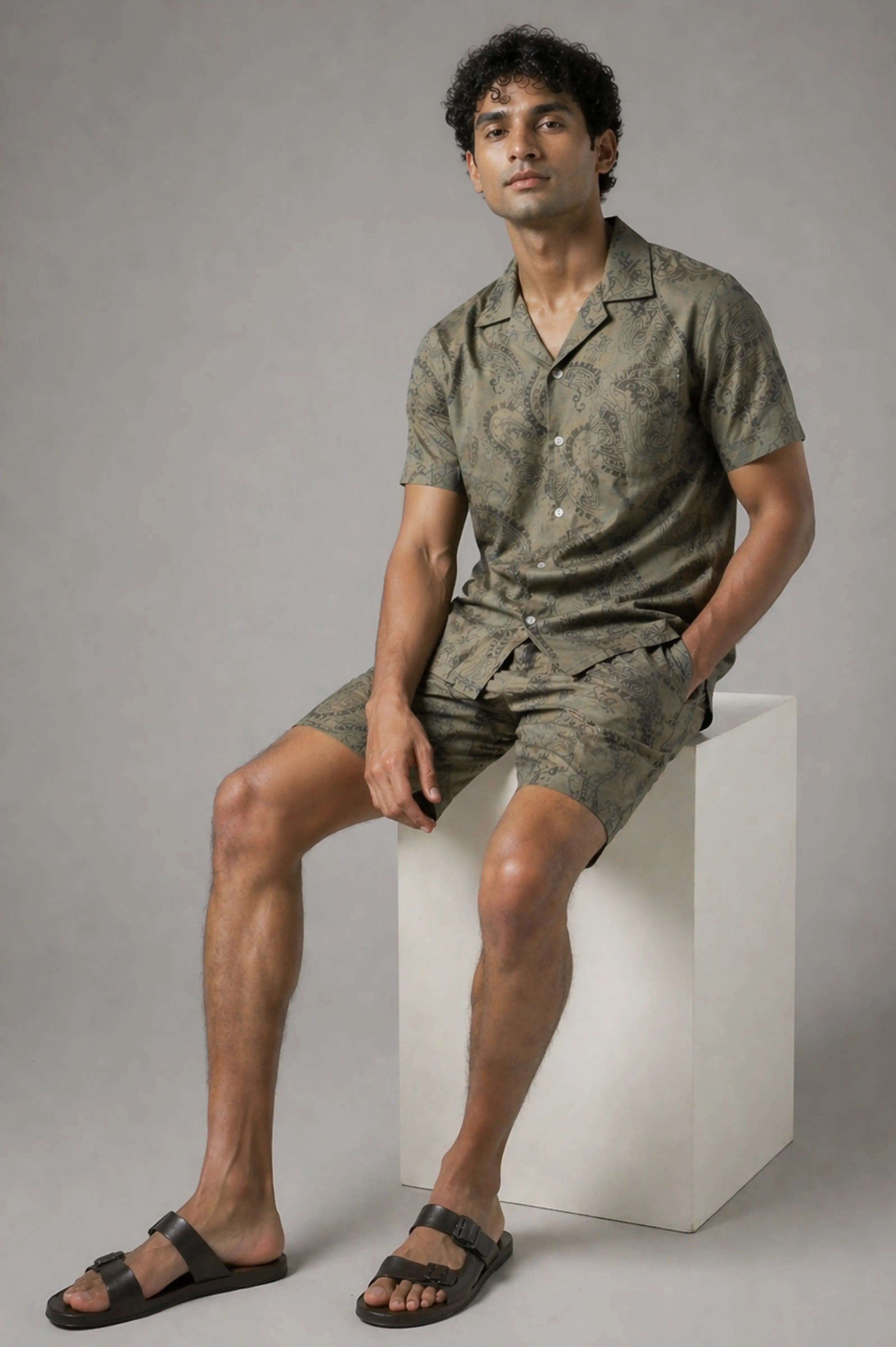 Man wearing a green patterned outfit sitting on a white block against a gray background
