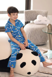 Child wearing blue pajamas with a car pattern. Sitting on a panda pillow in a living room.