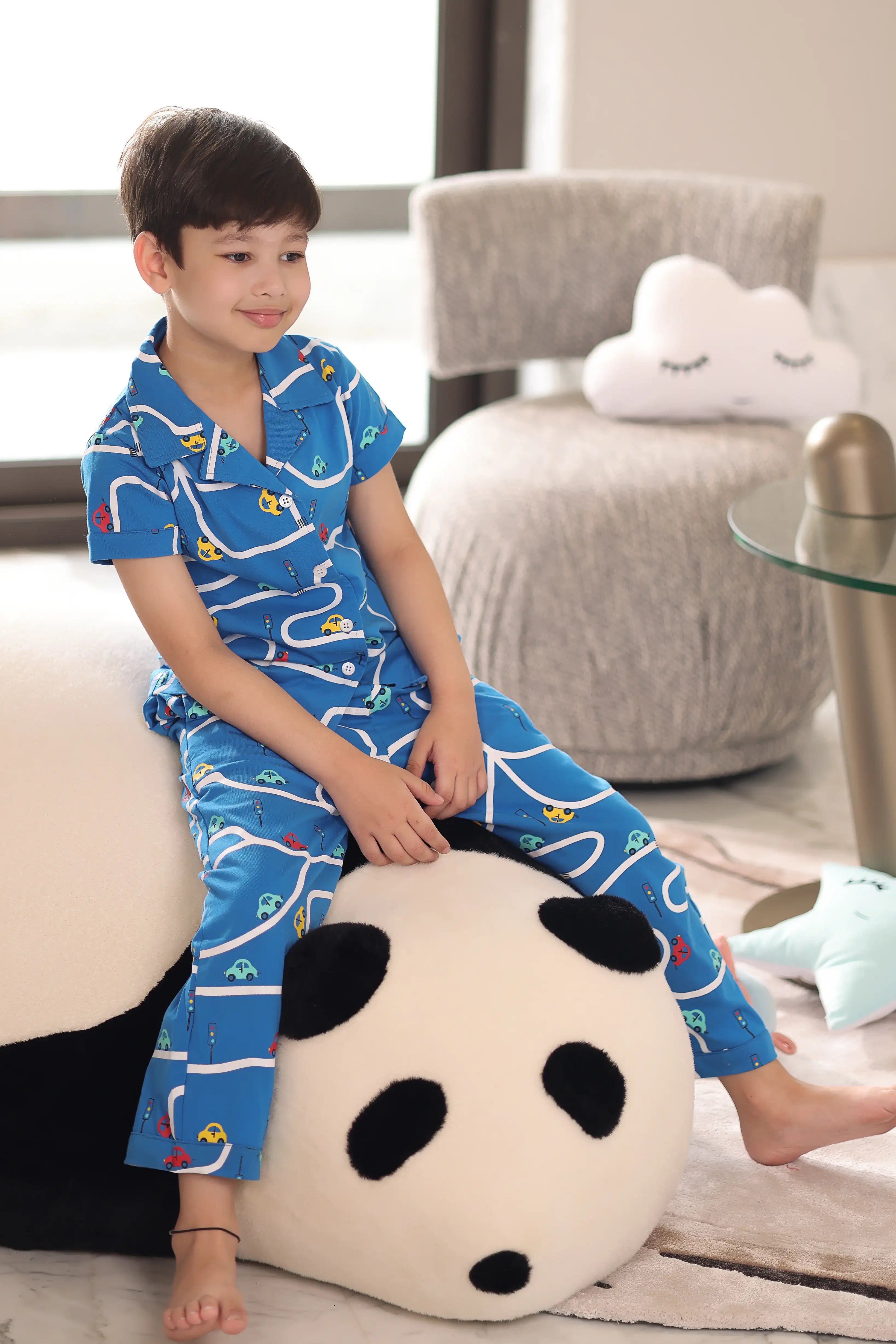Child wearing blue pajamas with a car pattern. Sitting on a panda pillow in a living room.