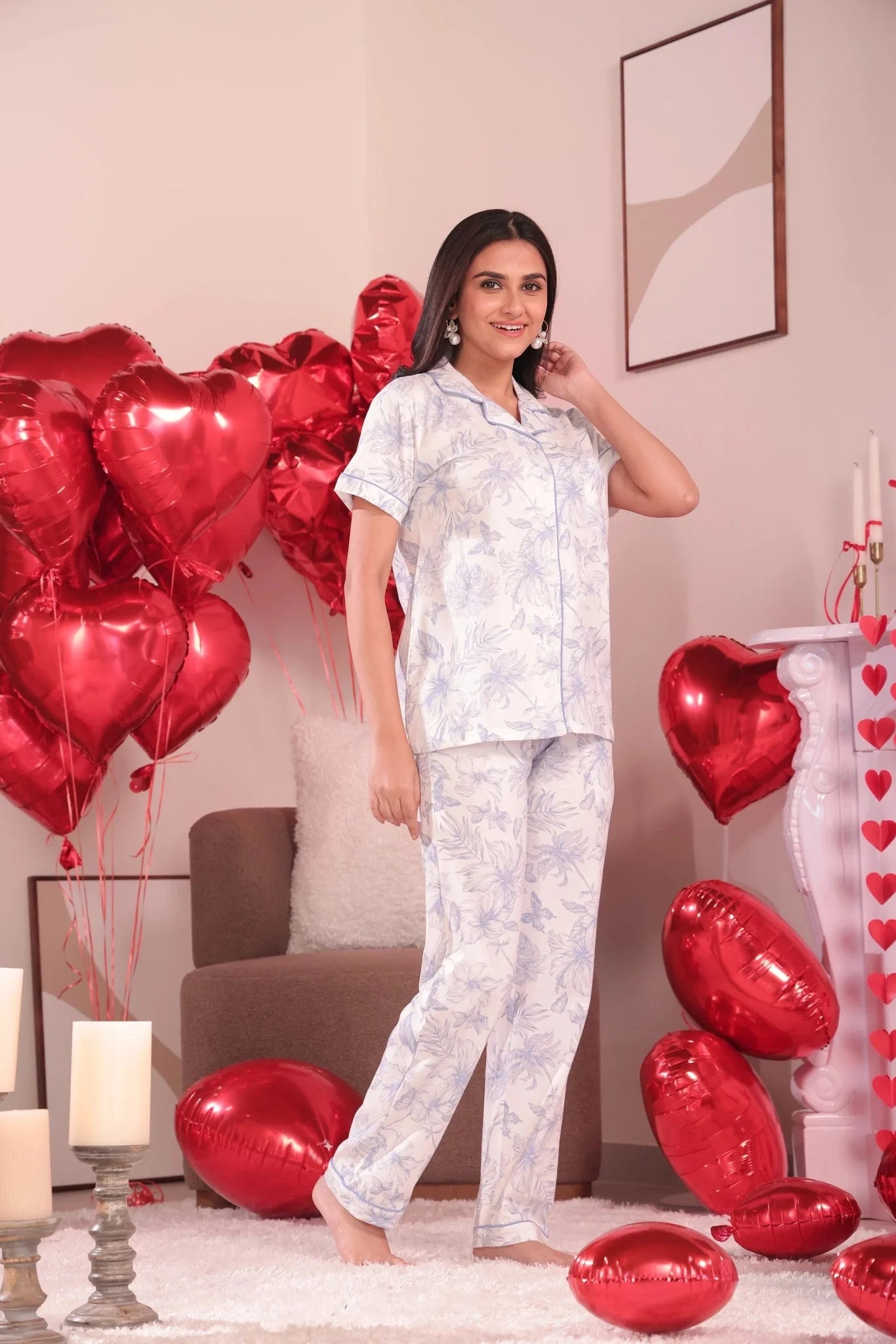 Woman in light-colored pajama set standing in a room with red heart-shaped balloons and candles.