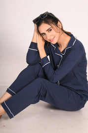 Young woman seated on the floor in a solid navy 100% cotton full sleeve nightsuit by NapStory, smiling warmly with her hands resting on her head.