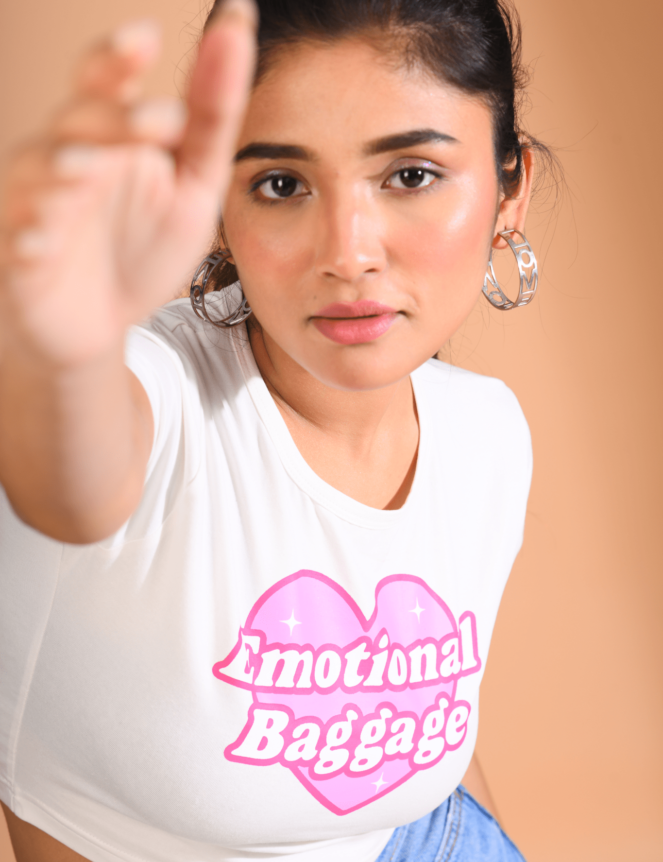 Woman wearing a white Baby Tee with a pink heart-shaped "Emotional Baggage" print, reaching forward with a focused expression.