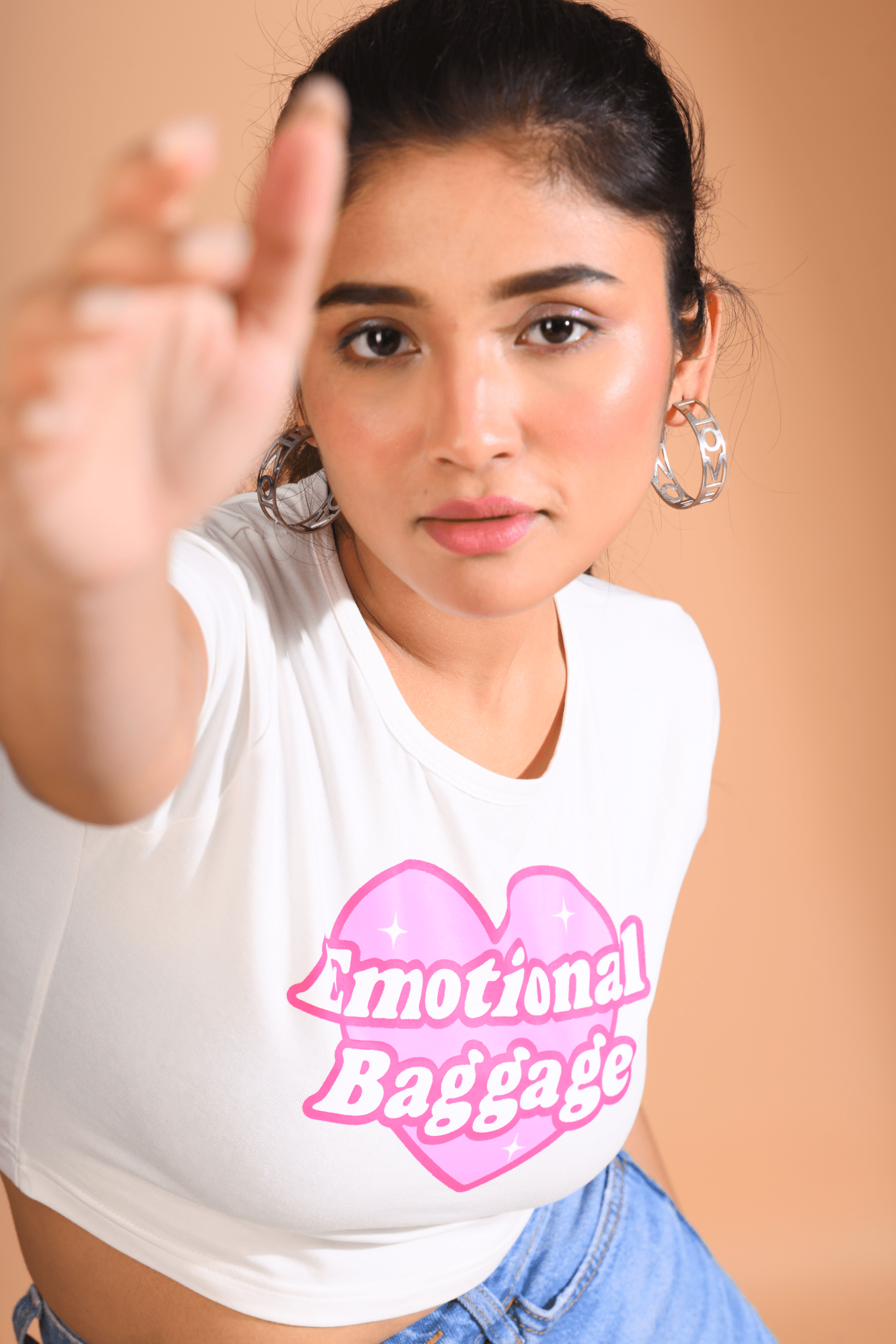 Woman wearing a white Baby Tee with a pink heart-shaped "Emotional Baggage" print, reaching forward with a focused expression.