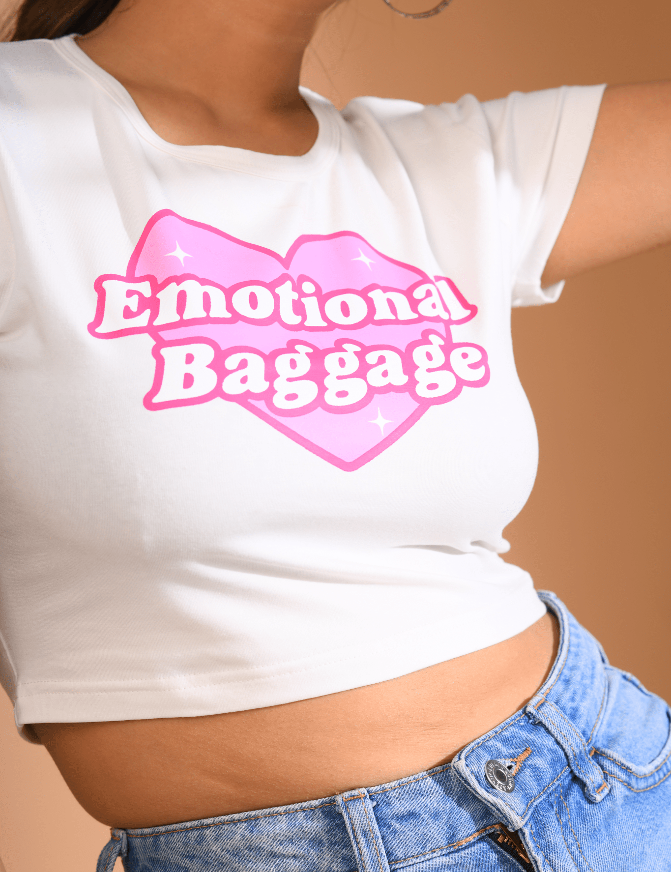 Close-up of a white Baby Tee with a pink heart-shaped "Emotional Baggage" print, paired with high-waisted blue jeans.