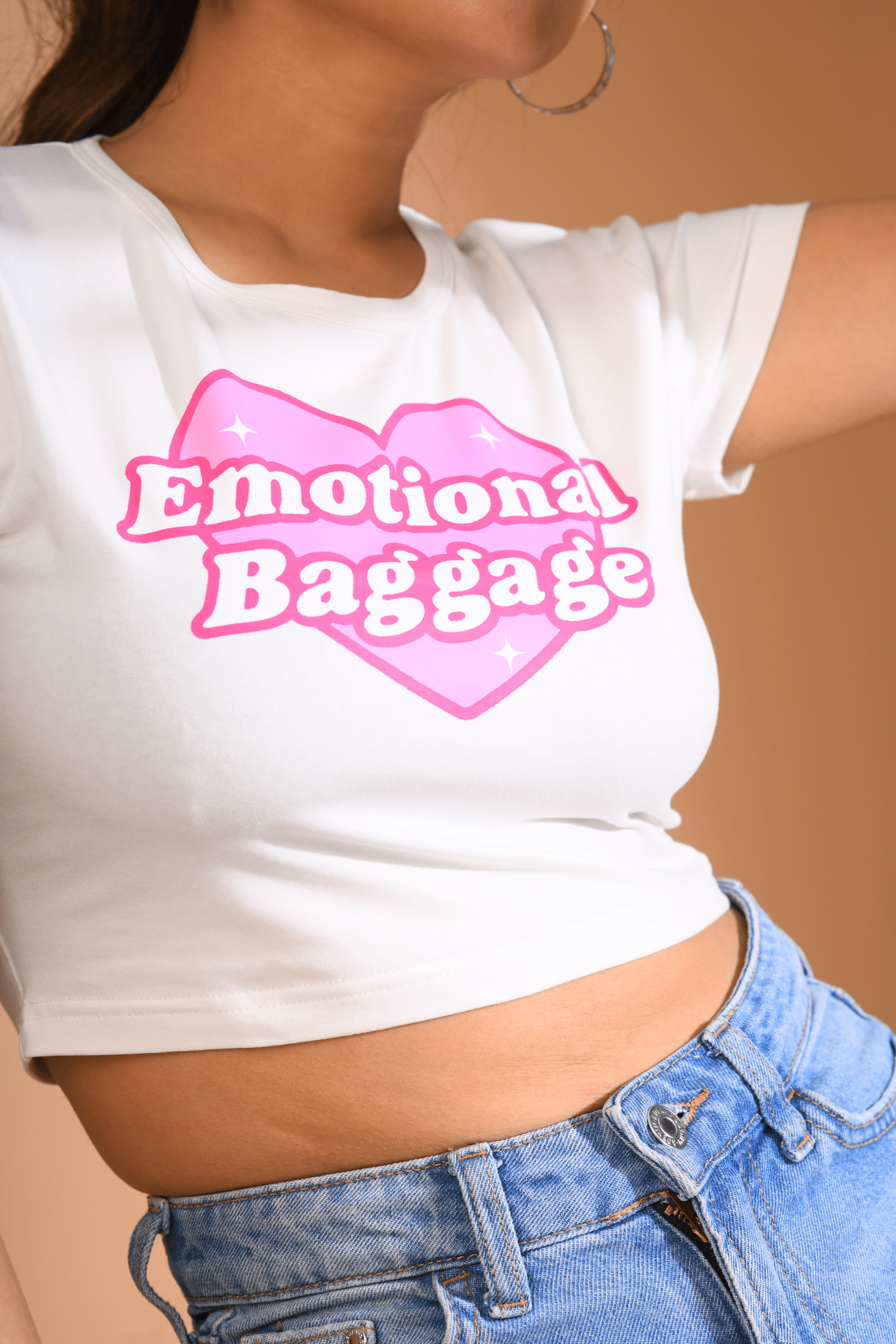 Close-up of a white Baby Tee with a pink heart-shaped "Emotional Baggage" print, paired with high-waisted blue jeans.